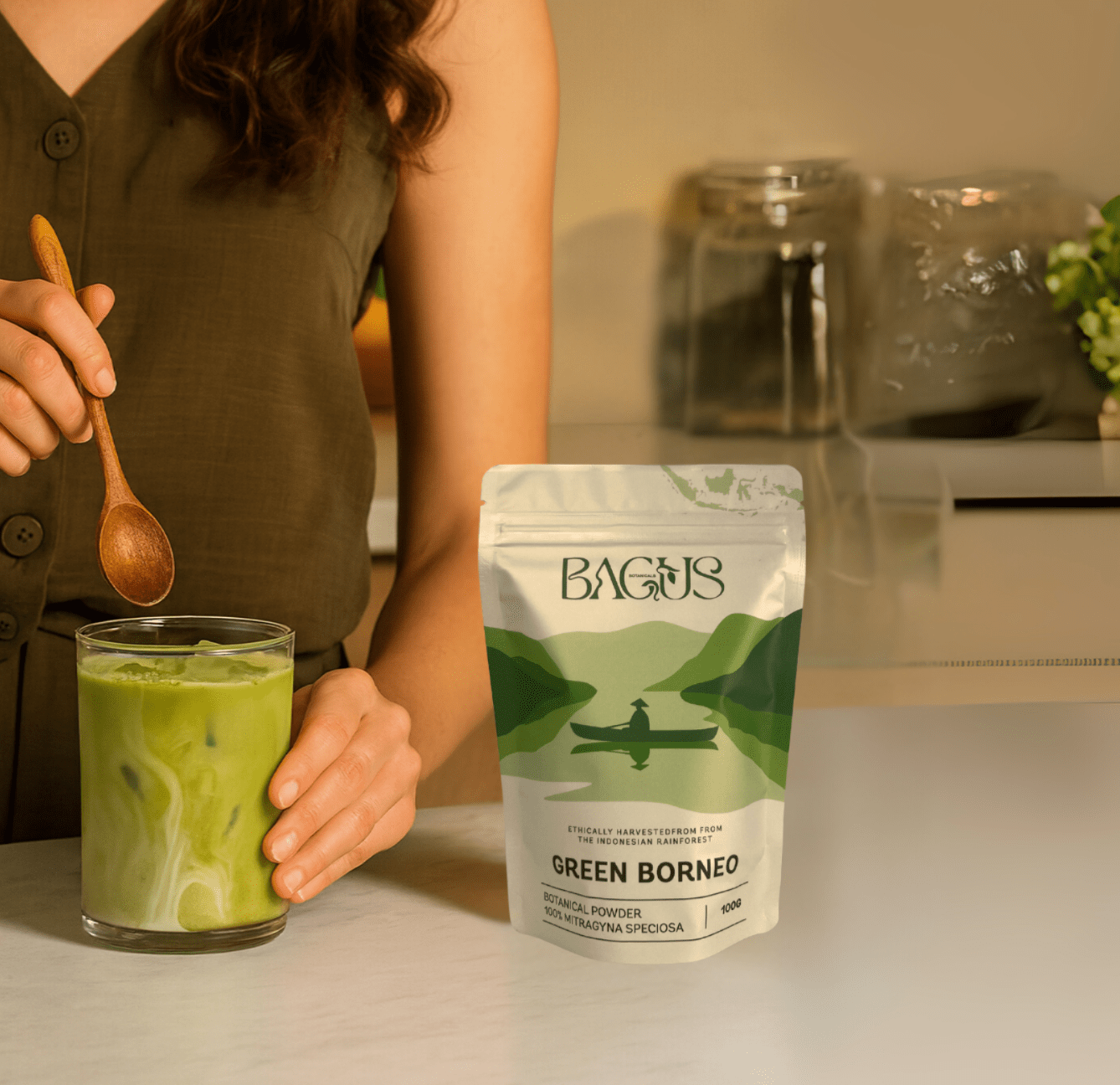 Woman stirring a glass of iced green drink made with Bagus Green Borneo kratom powder, product pouch on kitchen counter.